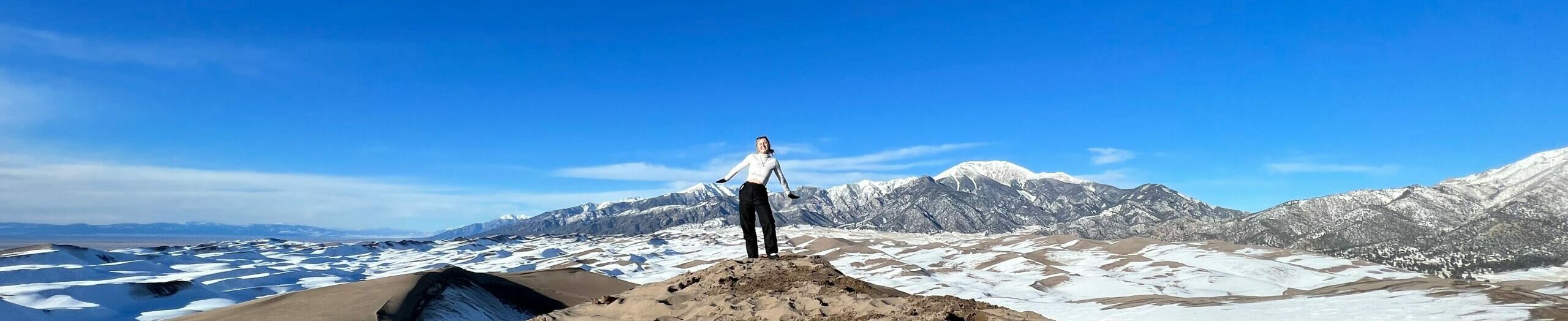 Jillian is standing on top of a sand dune at Great Sand Dunes National Park with mountains in the background.