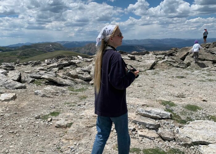 Jillian stands in the center holding a camera with mountain views stretching out around her.